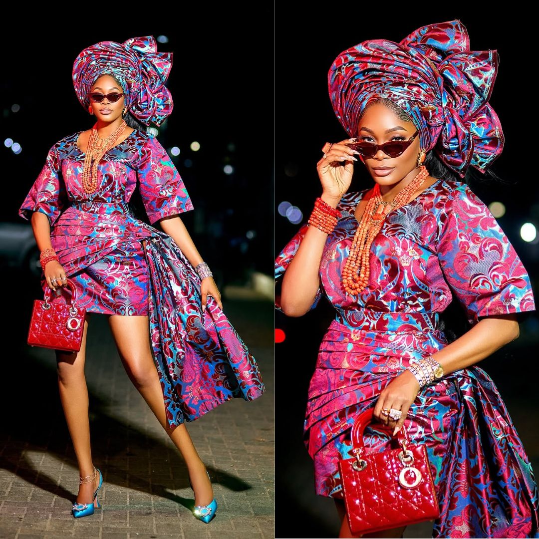 Media personality Kiekie posing in a multi-coloured silk brocade Oleku outfit featuring an asymmetrical mini wrapper with a dramatic side train, a matching voluminous gele, and coral beads.