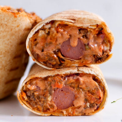 Two halves of a Nigerian beef shawarma wrap by Kikifoodies stacked vertically, showcasing a hotdog embedded in the centre of the juicy filling.