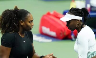 Tennis stars Serena Williams in a black athletic dress and Venus Williams in a white and green outfit shaking hands on a hard tennis court.