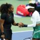 Tennis stars Serena Williams in a black athletic dress and Venus Williams in a white and green outfit shaking hands on a hard tennis court.
