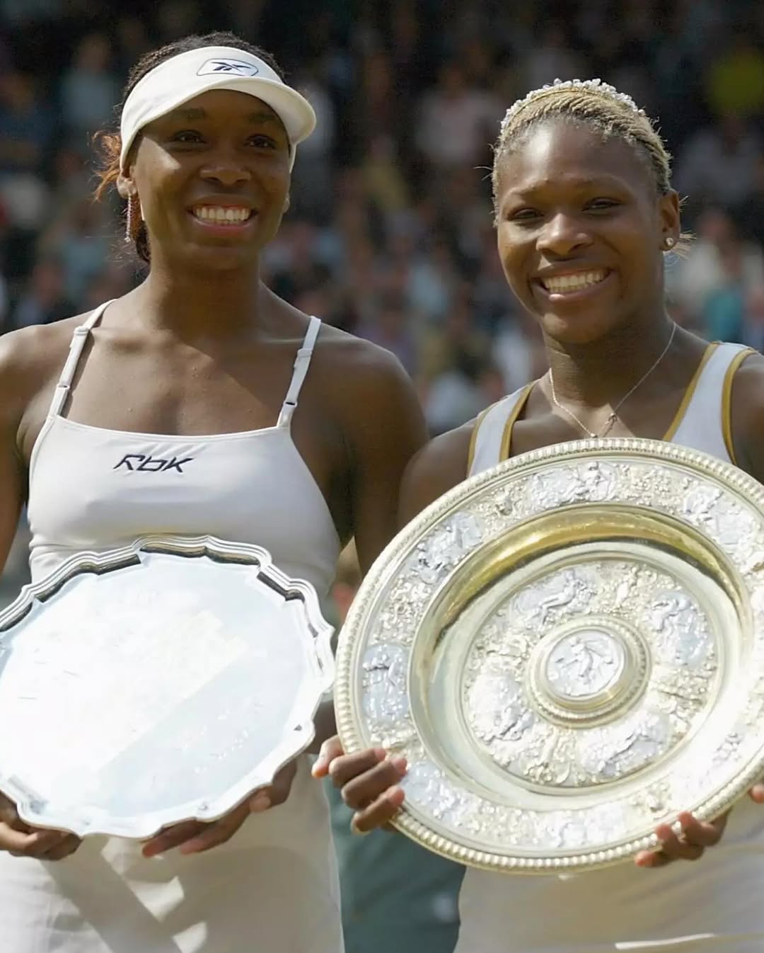 A young Venus and Serena Williams smiling broadly while holding up their silver Wimbledon singles and doubles championship trophies.