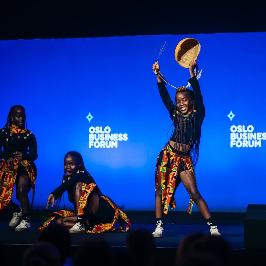 A member of the Ghetto Kids performing on stage at the Oslo Business Forum against a blue background, lifting a traditional calabash prop.