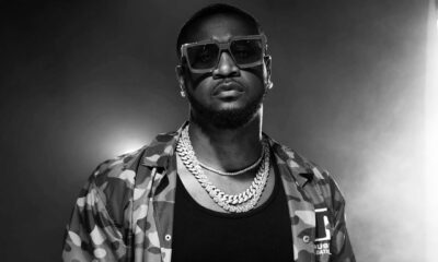 Peter Okoye, Mr P, wearing large sunglasses, diamond chain necklaces, a black vest, and an unbuttoned camouflage jacket.