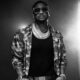 Peter Okoye, Mr P, wearing large sunglasses, diamond chain necklaces, a black vest, and an unbuttoned camouflage jacket.