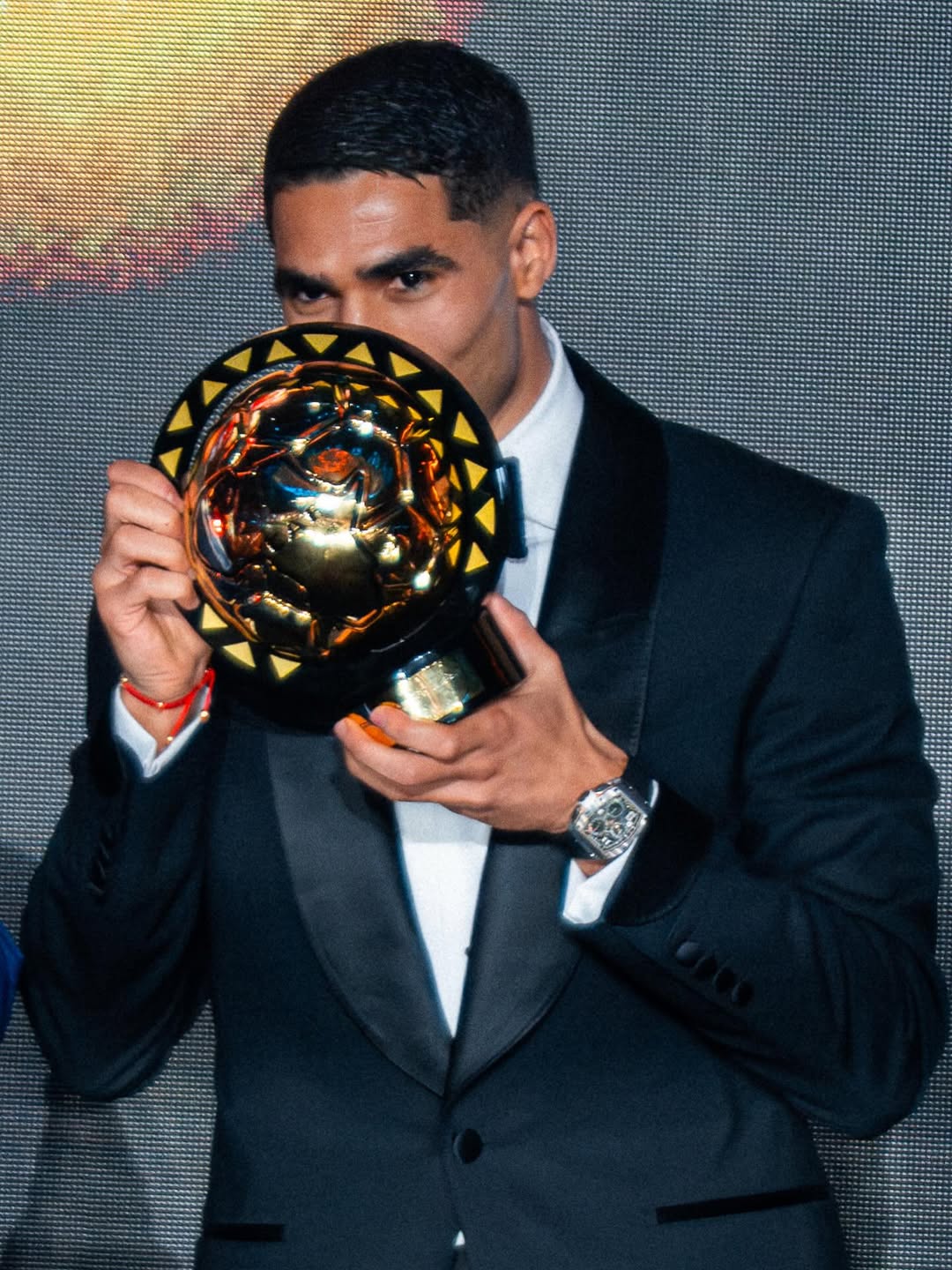 Morocco national football team captain Achraf Hakimi wearing a black tuxedo and holding the gold CAF Player of the Year trophy at an awards ceremony.