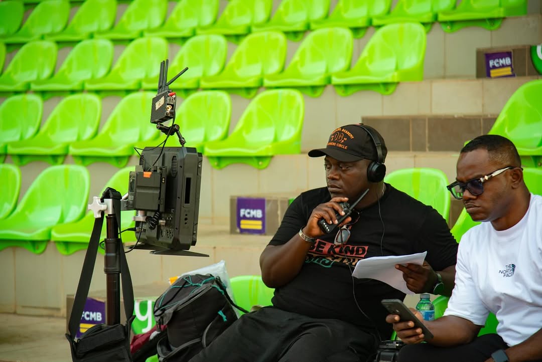 Nollywood director Adeoluwa Owu seated with headphones and a walkie-talkie looking at a camera monitor on the green-seated stadium set of Iwe Ala.