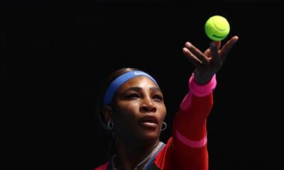 Serena Williams tossing a tennis ball into the air to serve on court, wearing a black, pink, and red Nike bodysuit.