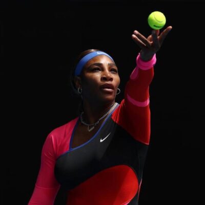 Serena Williams tossing a tennis ball into the air to serve on court, wearing a black, pink, and red Nike bodysuit.