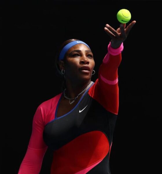 Serena Williams tossing a tennis ball into the air to serve on court, wearing a black, pink, and red Nike bodysuit.