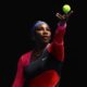 Serena Williams tossing a tennis ball into the air to serve on court, wearing a black, pink, and red Nike bodysuit.