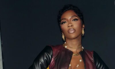 Singer Tiwa Savage poses with hands on her hips wearing a leather crop jacket, yellow top, and layered gold chain belts.