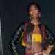 Singer Tiwa Savage poses with hands on her hips wearing a leather crop jacket, yellow top, and layered gold chain belts.