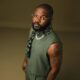 A high-angle portrait of Nigerian artist Falz wearing a sleeveless olive green top and an embellished scarf, highlighting his distinct arm tattoos.
