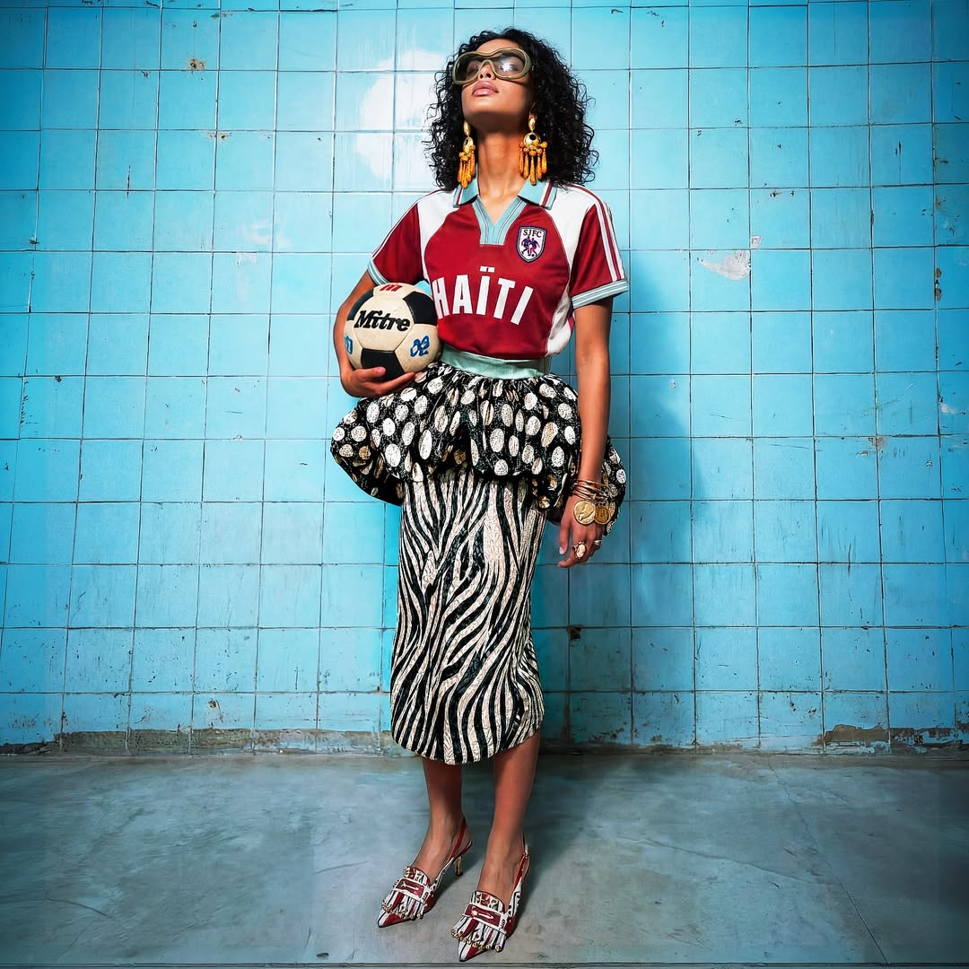 A model standing against a blue tiled wall holding a football, wearing Stella Jean's burgundy L'Haitiana Haiti jersey paired with a dotted black-and-white bubble peplum and zebra print pencil skirt.