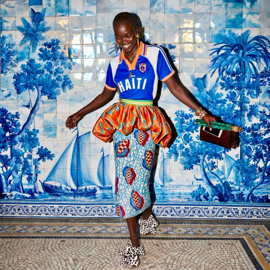 A model smiling against a blue and white mosaic wall, wearing the royal blue L'Haitiana Haiti football jersey with a bright orange gathered bubble waist and metallic blue and gold brocade midi skirt.