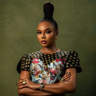 Close-up of Nancy Isime with crossed arms, showcasing her regal suku braided topknot, metallic almond-shaped nails, gold rings, and luxury wristwatch.
