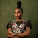 Close-up of Nancy Isime with crossed arms, showcasing her regal suku braided topknot, metallic almond-shaped nails, gold rings, and luxury wristwatch.