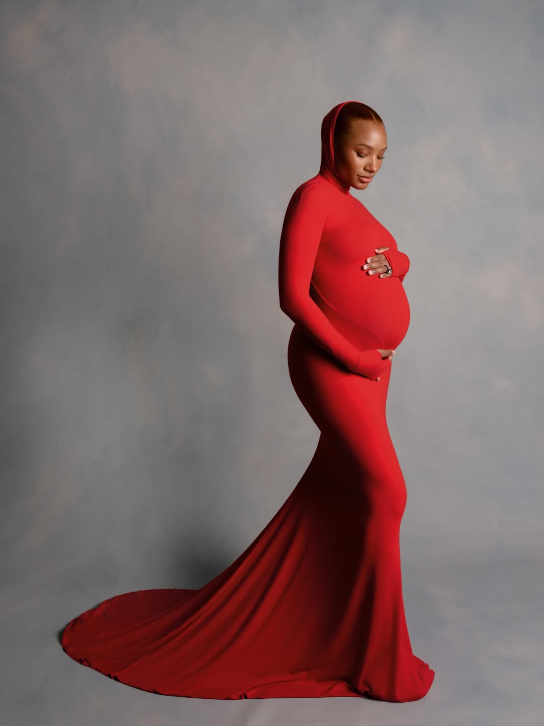 A formal studio portrait of Temi Otedola in profile wearing a minimalist, form-fitting red jersey maternity gown with an integrated hood and pooling train.