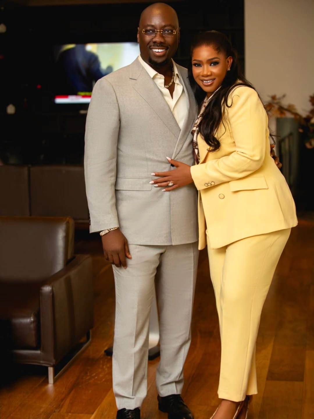 Ebele Iyiegbu placing a hand on her husband Obi Cubana's waist as they pose together in their stone grey and butter yellow tailored suits.