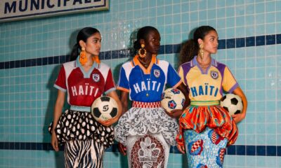 Three models posing by an indoor swimming pool holding footballs, showcasing Stella Jean's L'Haitiana special-edition hand-stitched jerseys for the Haiti national football club in burgundy, blue, and yellow.