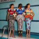 Three models posing by an indoor swimming pool holding footballs, showcasing Stella Jean's L'Haitiana special-edition hand-stitched jerseys for the Haiti national football club in burgundy, blue, and yellow.