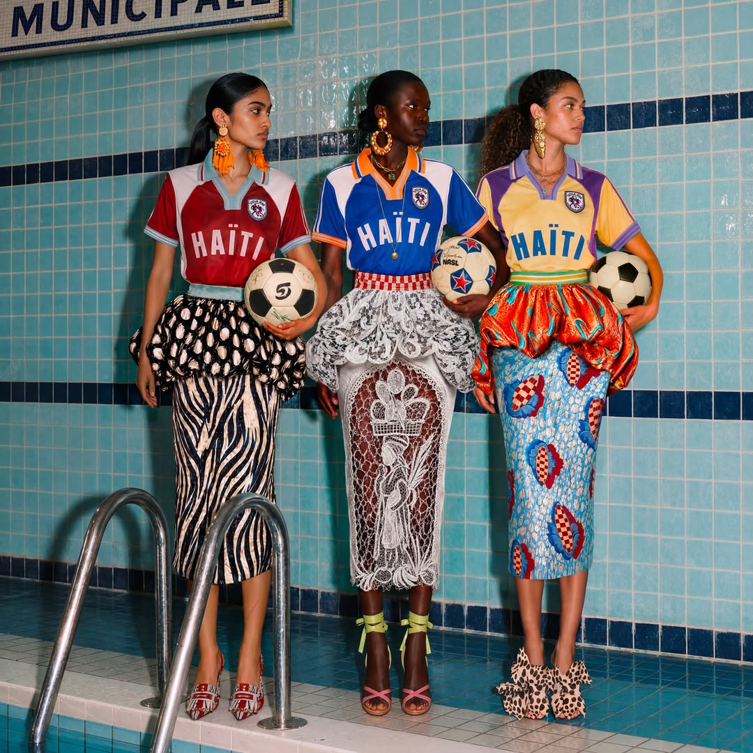 Three models posing by an indoor swimming pool holding footballs, showcasing Stella Jean's L'Haitiana special-edition hand-stitched jerseys for the Haiti national football club in burgundy, blue, and yellow.