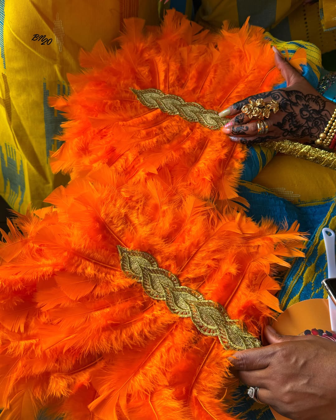 A close-up of hands adorned with intricate black henna tattoos and large gold rings holding vibrant orange feather fans at Ojude Oba.