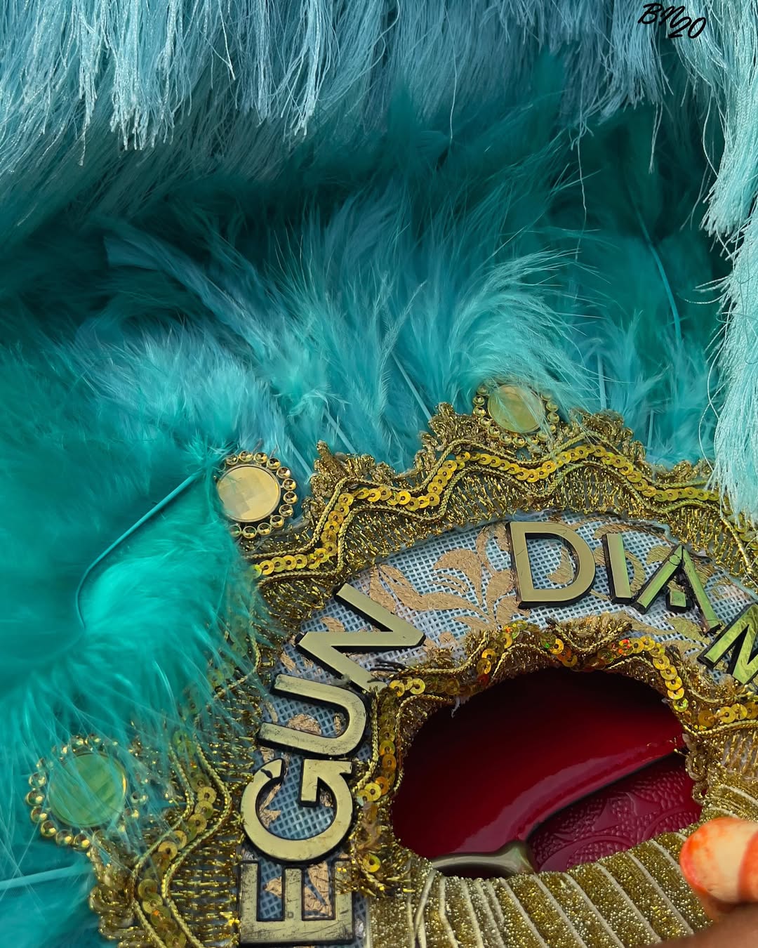 An ornate turquoise blue feather hand fan with gold lace trim, gold lettering spelling "EGUN", and mirrors at an Ijebu cultural event.