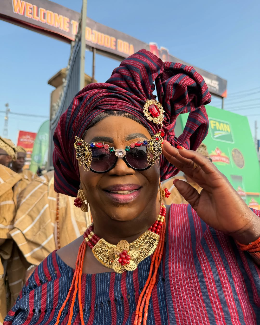 A close-up of a woman at Ojude Oba 2026 wearing dark sunglasses adorned with ornate gold butterfly wings and multi-coloured jewels, paired with a wine and navy Aso Oke.