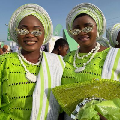 Two Ijebu-Ode women smiling at the Ojude Oba Festival wearing matching lime green buba, white and green gele, and stone-embellished sunglasses.