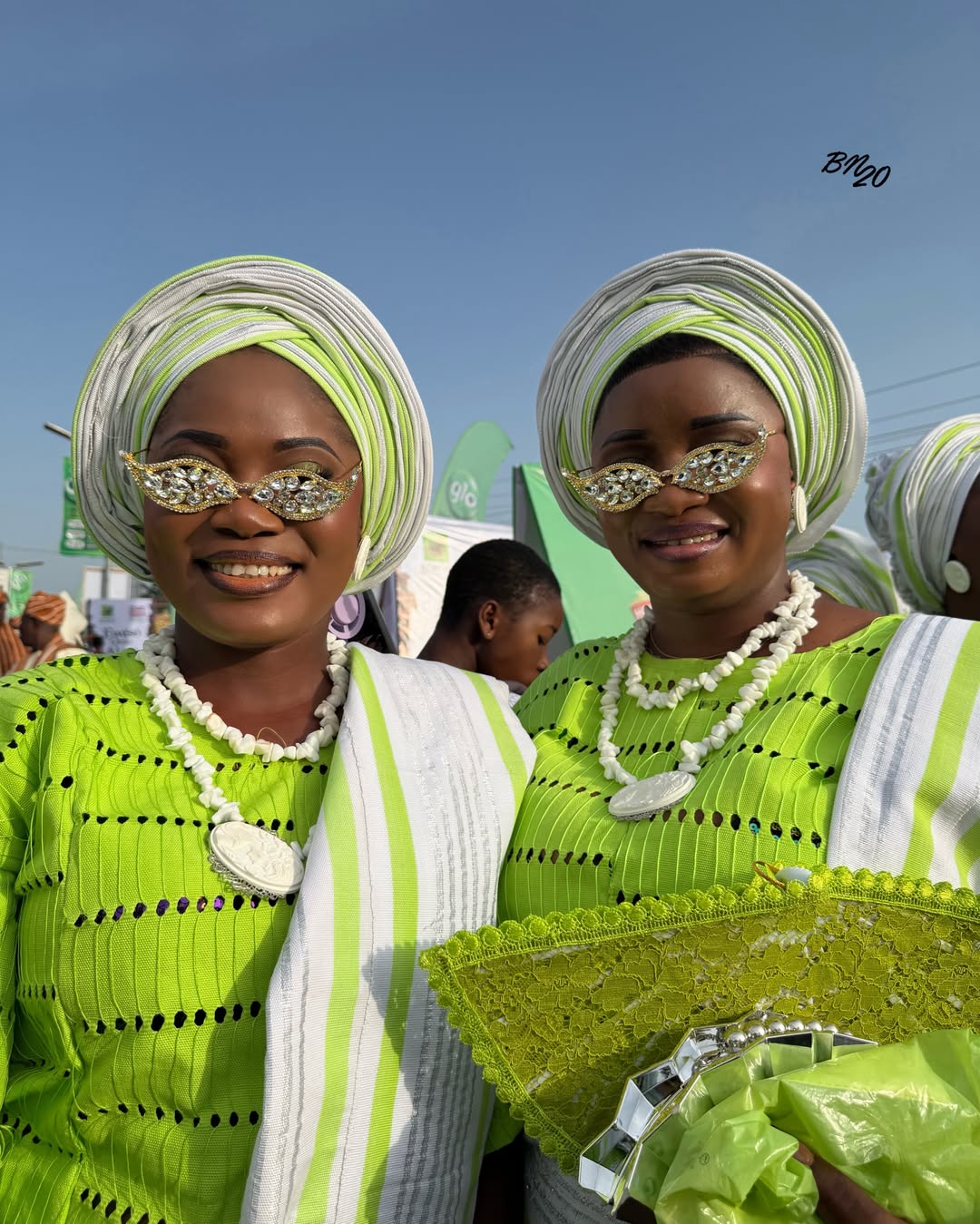 Two Ijebu-Ode women smiling at the Ojude Oba Festival wearing matching lime green buba, white and green gele, and stone-embellished sunglasses.