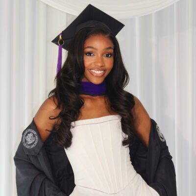 Chioma Amadi smiling in a white strapless dress with her black Savannah College of Art and Design graduation gown draped loosely over her arms.