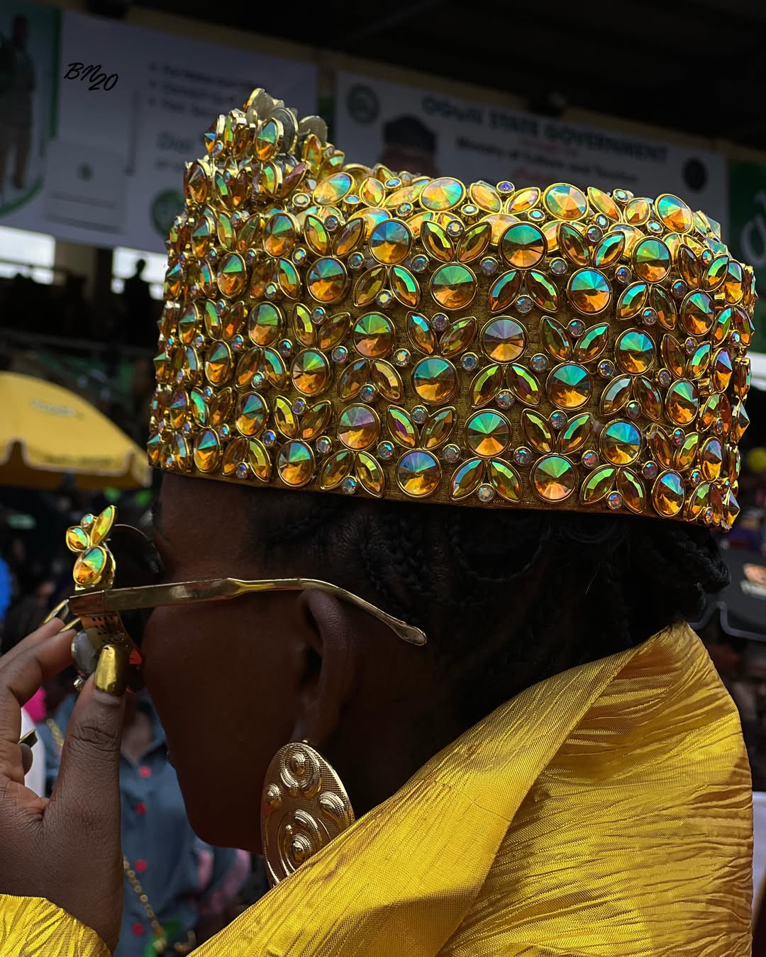 A woman profile view wearing an iridescent orange and gold embellished hat with gold sunglasses and heavy gold earrings at the festival.