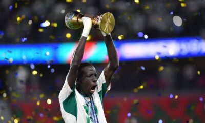A Senegal youth football player shouting in celebration while lifting the golden U17 AFCON trophy amidst falling gold confetti at the stadium.