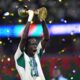 A Senegal youth football player shouting in celebration while lifting the golden U17 AFCON trophy amidst falling gold confetti at the stadium.