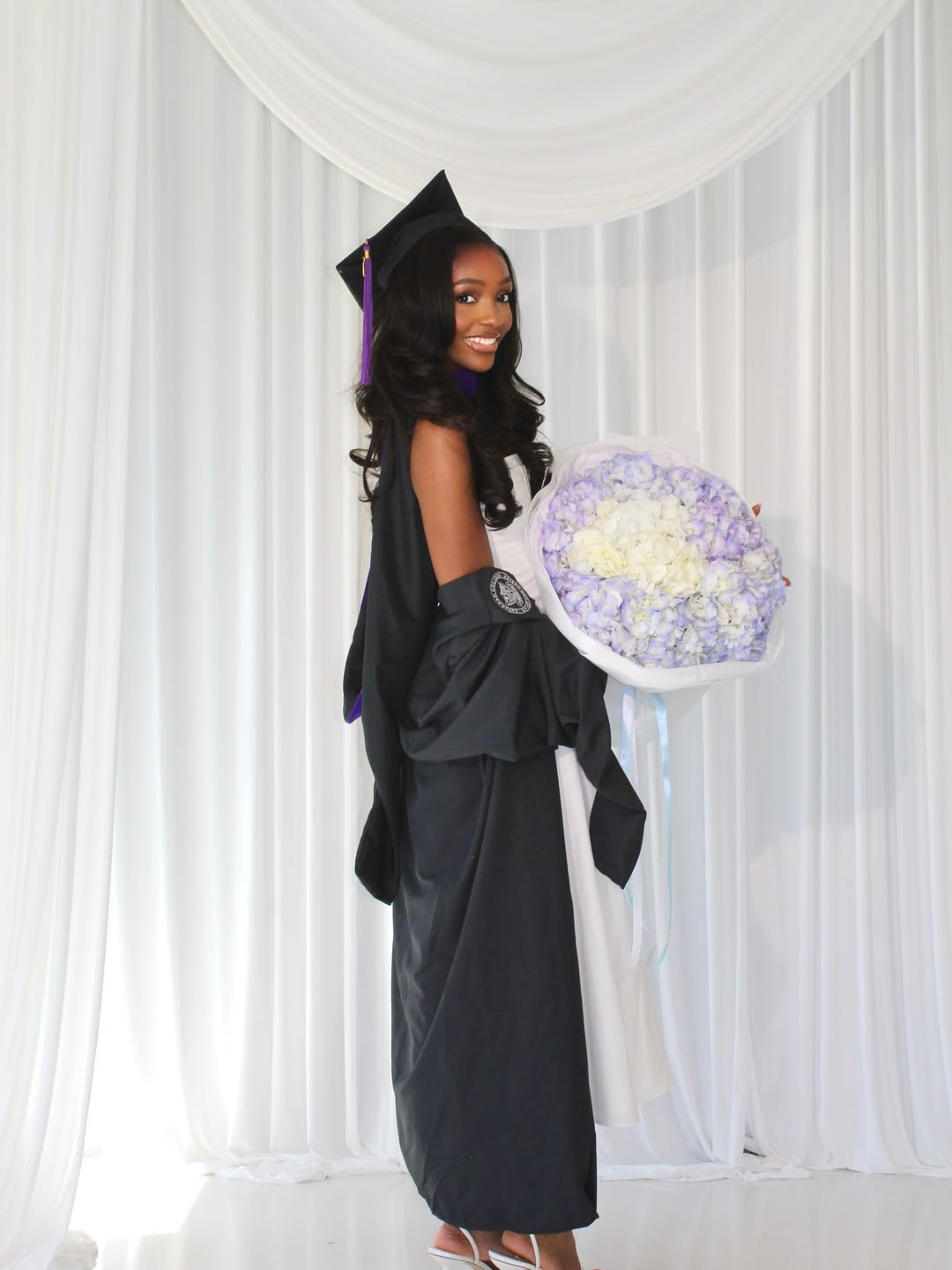 Former Miss Nigeria Chioma Amadi smiling in a black graduation gown and cap while holding a large bouquet of purple and white hydrangeas.