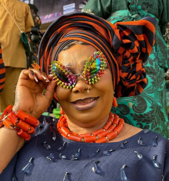 A smiling attendee at Ojude Oba 2026 holding multi-coloured iridescent sunglasses bordered with metallic spikes, paired with a navy blue outfit and heavy orange coral beads.