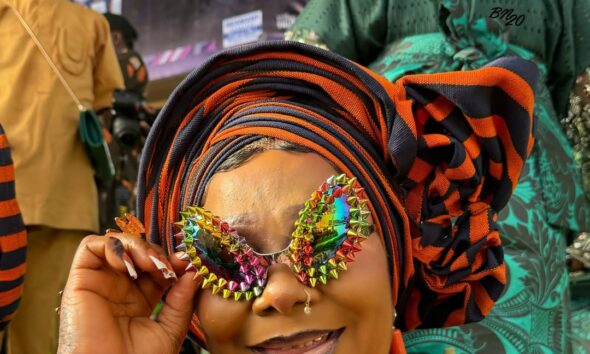 A smiling attendee at Ojude Oba 2026 holding multi-coloured iridescent sunglasses bordered with metallic spikes, paired with a navy blue outfit and heavy orange coral beads.