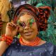 A smiling attendee at Ojude Oba 2026 holding multi-coloured iridescent sunglasses bordered with metallic spikes, paired with a navy blue outfit and heavy orange coral beads.