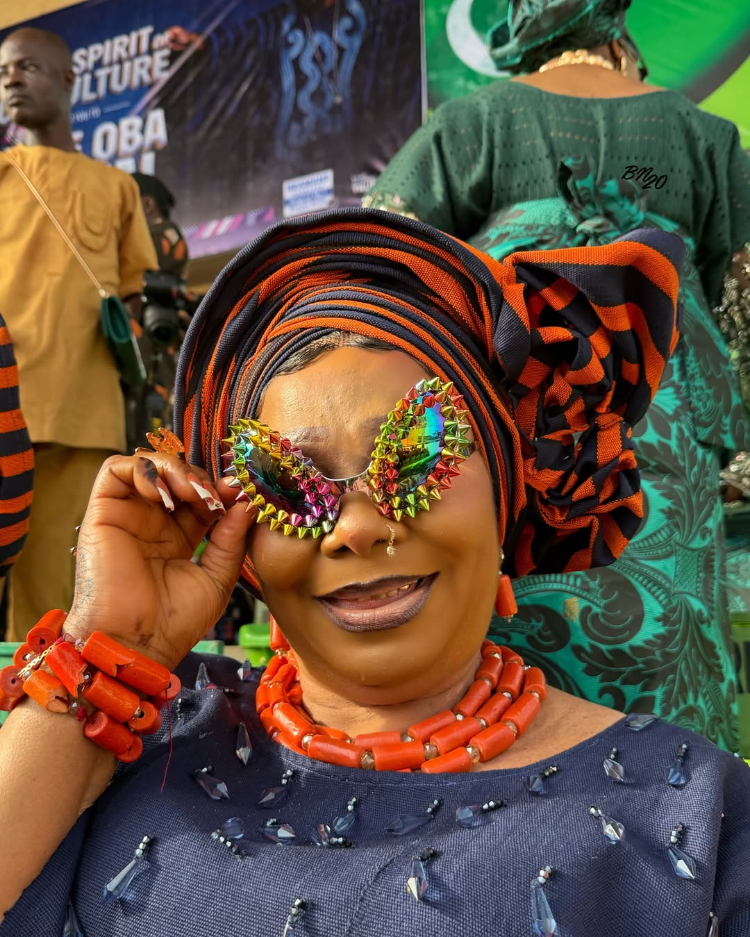 A smiling attendee at Ojude Oba 2026 holding multi-coloured iridescent sunglasses bordered with metallic spikes, paired with a navy blue outfit and heavy orange coral beads.