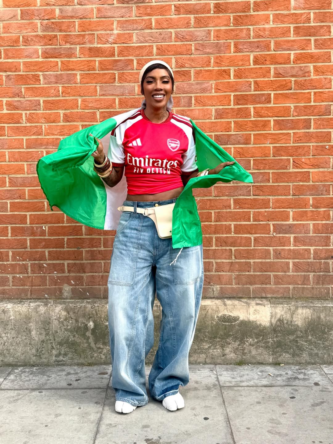 Afrobeats artist Tiwa Savage smiling while holding open the green and white Nigerian flag over her shoulders during the Arsenal trophy celebration in London.