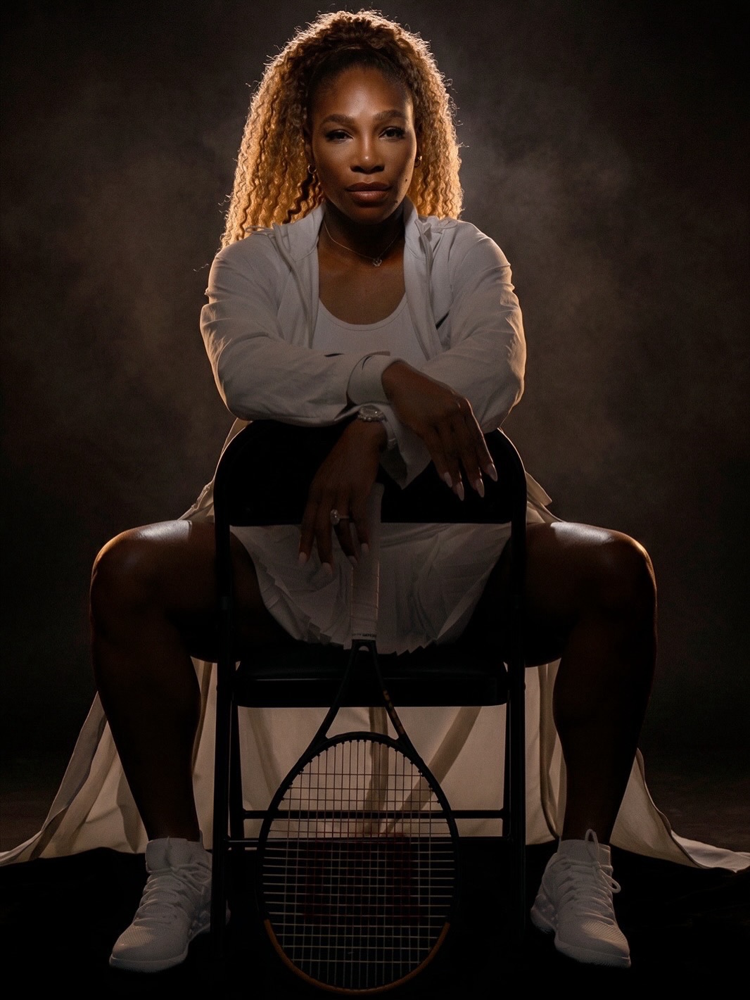 A dramatic, low-lit studio portrait of Serena Williams sitting on a folding chair in a white tennis outfit, leaning forward over a tennis racket.