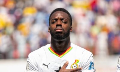 Ghana national football team forward Iñaki Williams standing on the pitch with his eyes closed and his right hand placed over the Black Star crest on his white jersey during a match.