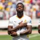 Ghana national football team forward Iñaki Williams standing on the pitch with his eyes closed and his right hand placed over the Black Star crest on his white jersey during a match.