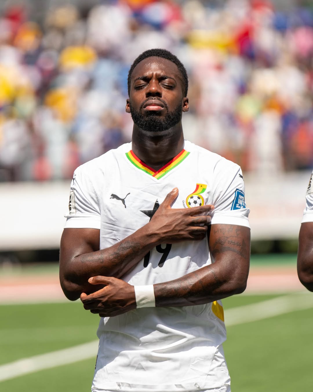 Ghana national football team forward Iñaki Williams standing on the pitch with his eyes closed and his right hand placed over the Black Star crest on his white jersey during a match.