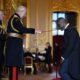 Idris Elba kneeling on a red velvet stool as King Charles III touches his shoulder with a ceremonial sword during his knighthood investiture ceremony inside Windsor Castle.