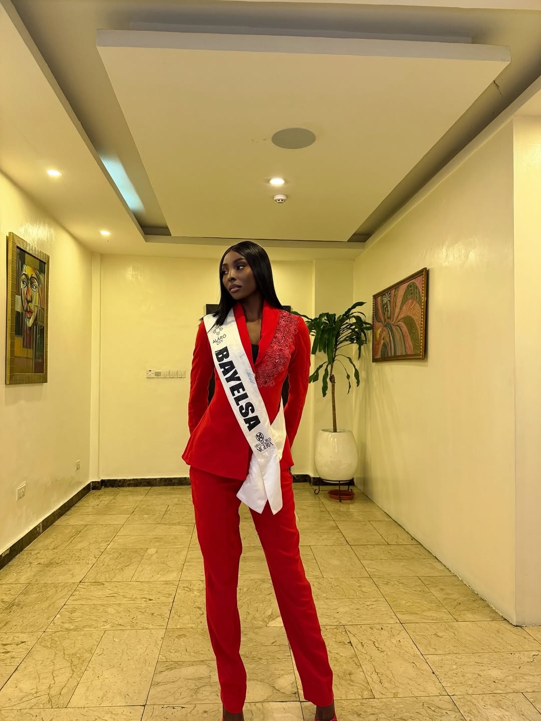 Miss World Nigeria contestant Tamunosoye Karibi George wearing her official white Bayelsa state sash over a red tailored pantsuit.