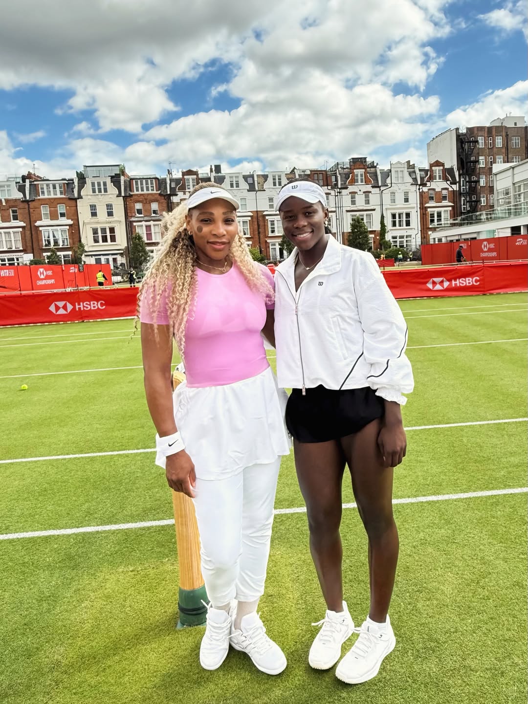 Serena Williams in a pink top and Victoria Mboko in a white jacket standing together on the grass courts at the HSBC Championships at Queen's Club in London.