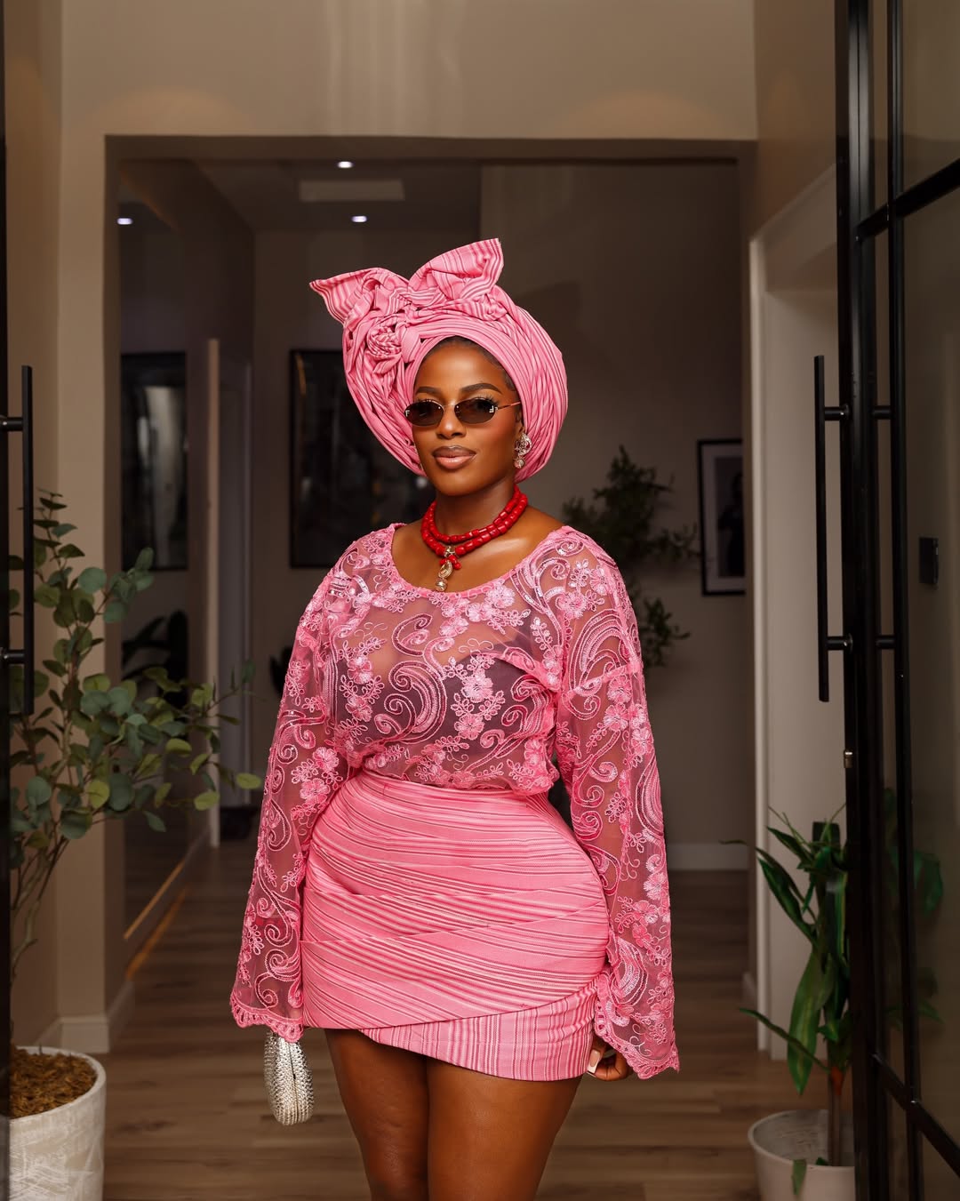 Hilda Baci posing in a doorway holding a silver clutch bag, showcasing an Emmanuel Goodnews styled look featuring a pink lace buba blouse and draped aso-oke mini skirt.