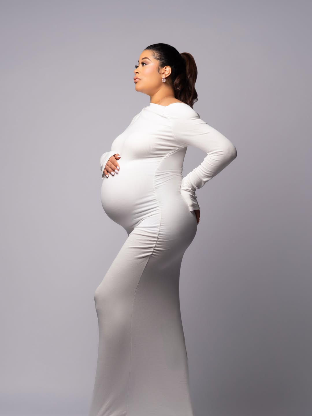 Side-profile maternity shoot of Nollywood actress Adunni Ade wearing a form-fitting white long-sleeved dress and cradling her baby bump.